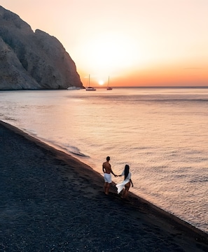 Escena de playa con una pareja tomados de la mano, un atardecer y barcos a lo lejos.