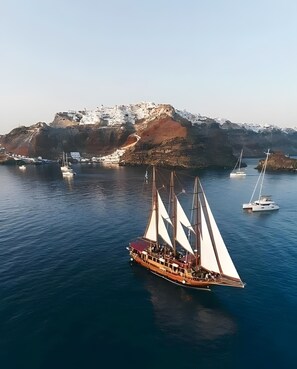 Um grande navio a vela com vários mastros e um convés vermelho, ancorado perto de uma costa rochosa, com alguns barcos menores ao fundo.