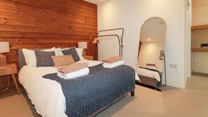 A bedroom with a wooden wall, a bed with a blue blanket, a bedside table with a lamp, and a mirror. - Teach Tra West (Perranporth)