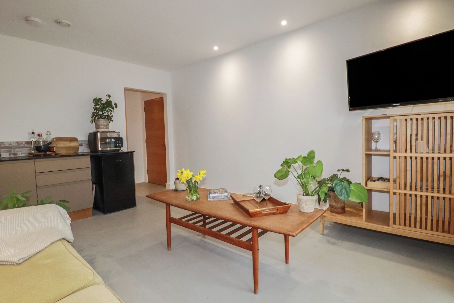 A modern living room with a wooden coffee table, a TV mounted on the wall, and a kitchen area with a microwave and a plant.