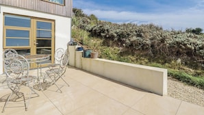 A modern building with a patio area featuring outdoor furniture and a view of a hillside. - Teach Tra West (Perranporth)
