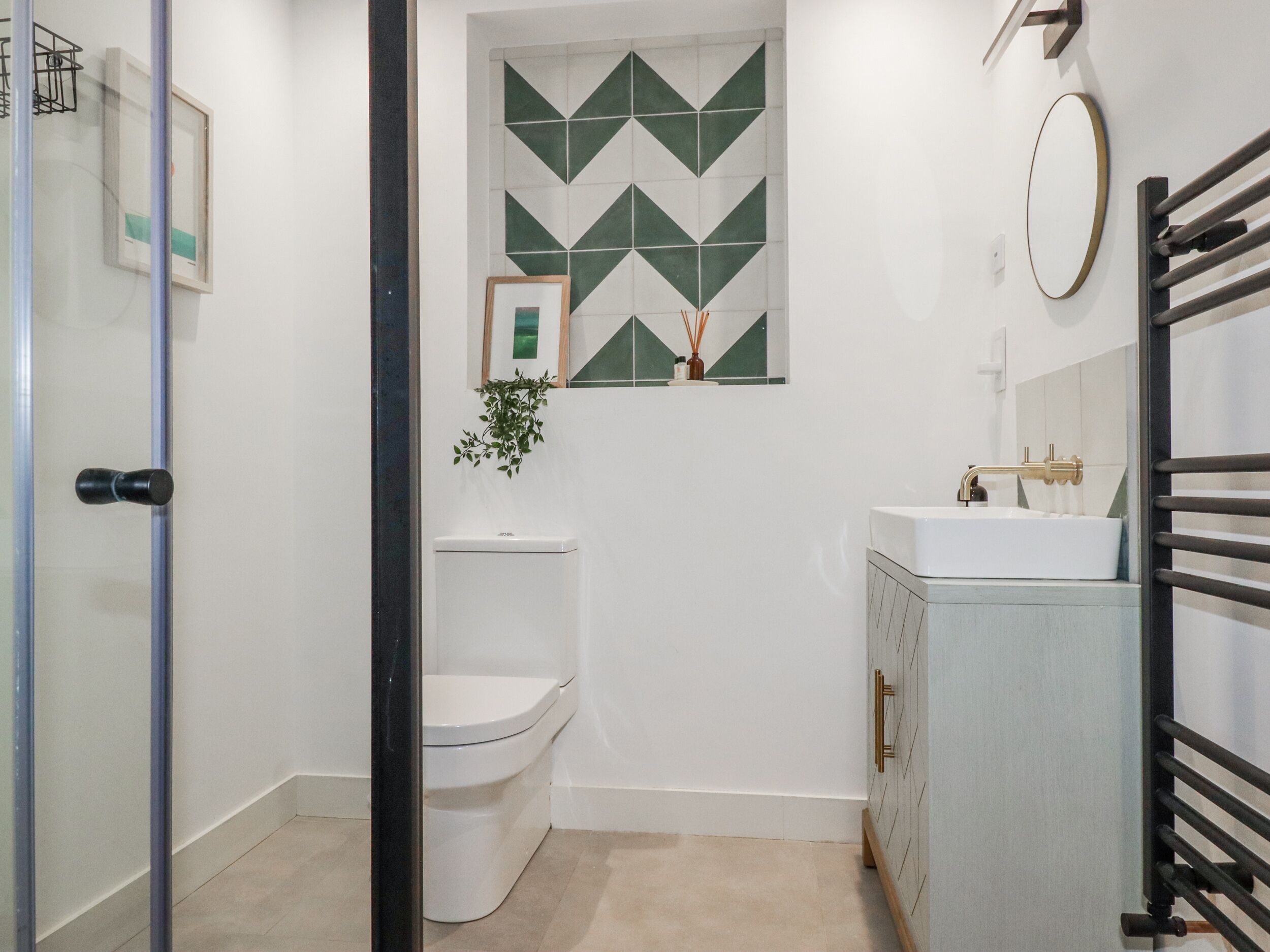 Ein kleines, modernes Badezimmer mit Toilette in Weiß, einem weißen Waschbecken mit goldfarbenem Wasserhahn und einem Spiegel darüber. Eine Wanddekoration mit geometrischen Mustern in Grün und Weiß befindet sich über der Toilette.