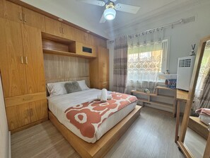 A bedroom with a bed, wooden wardrobe, ceiling fan, and a desk with a mirror.