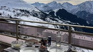 Een eettafel met borden, kommen en toevoegingen op een balkon met uitzicht op besneeuwde bergen.