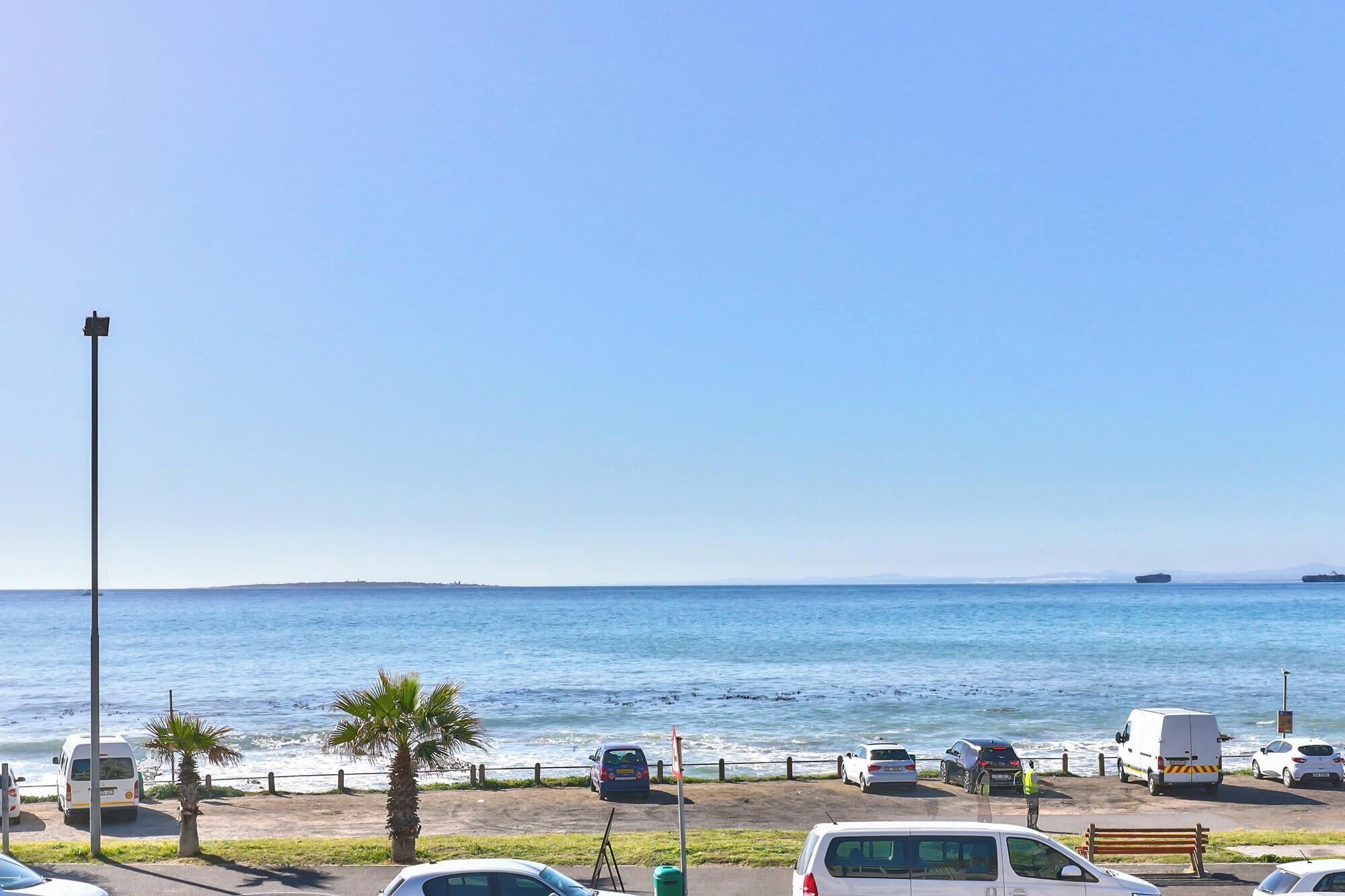 A parking area with vehicles, a palm tree, and a view of the sea.