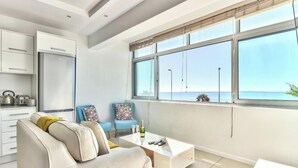 Un salon moderne comprenant un canapé, deux fauteuils, une table basse et une vue sur la mer grâce à de grandes fenêtres.