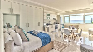 Une chambre moderne avec un grand lit, un coin salon avec une table et des chaises, une cuisine avec des meubles blancs et une vue sur la mer grâce à de grandes fenêtres.