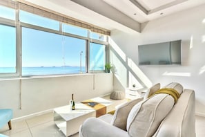 Ein modernes Wohnzimmer mit großem Fenster zum Meer, einem an der Wand montierten Flachbildfernseher und einer gemütlichen Sitzecke mit Couchtisch.