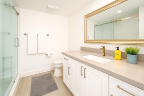 A modern bathroom with a glass shower enclosure, a toilet, a sink with a gold faucet, and a mirror with a gold frame.