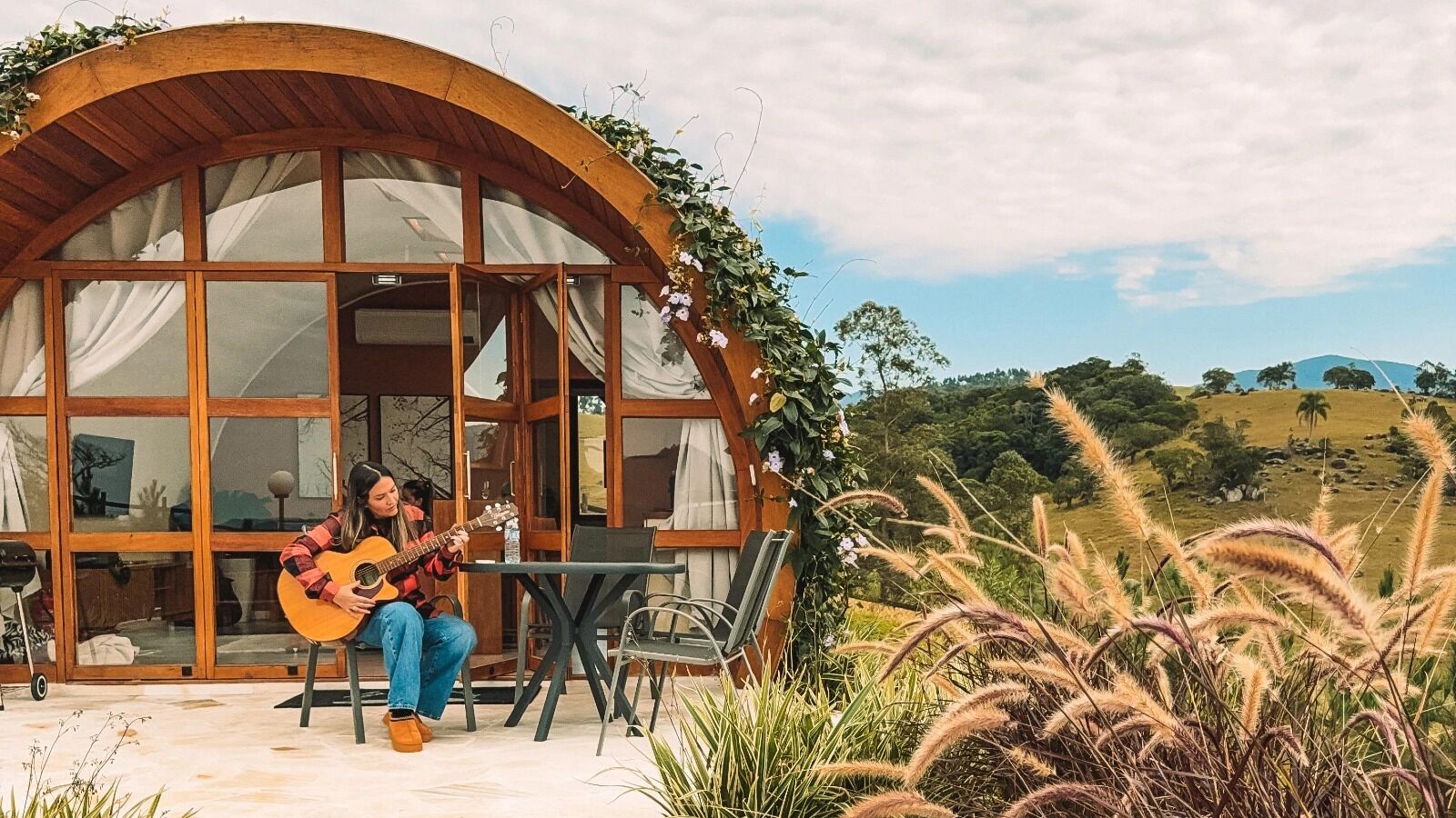 A person playing a guitar at a table outside a building with large windows and a wooden arch.