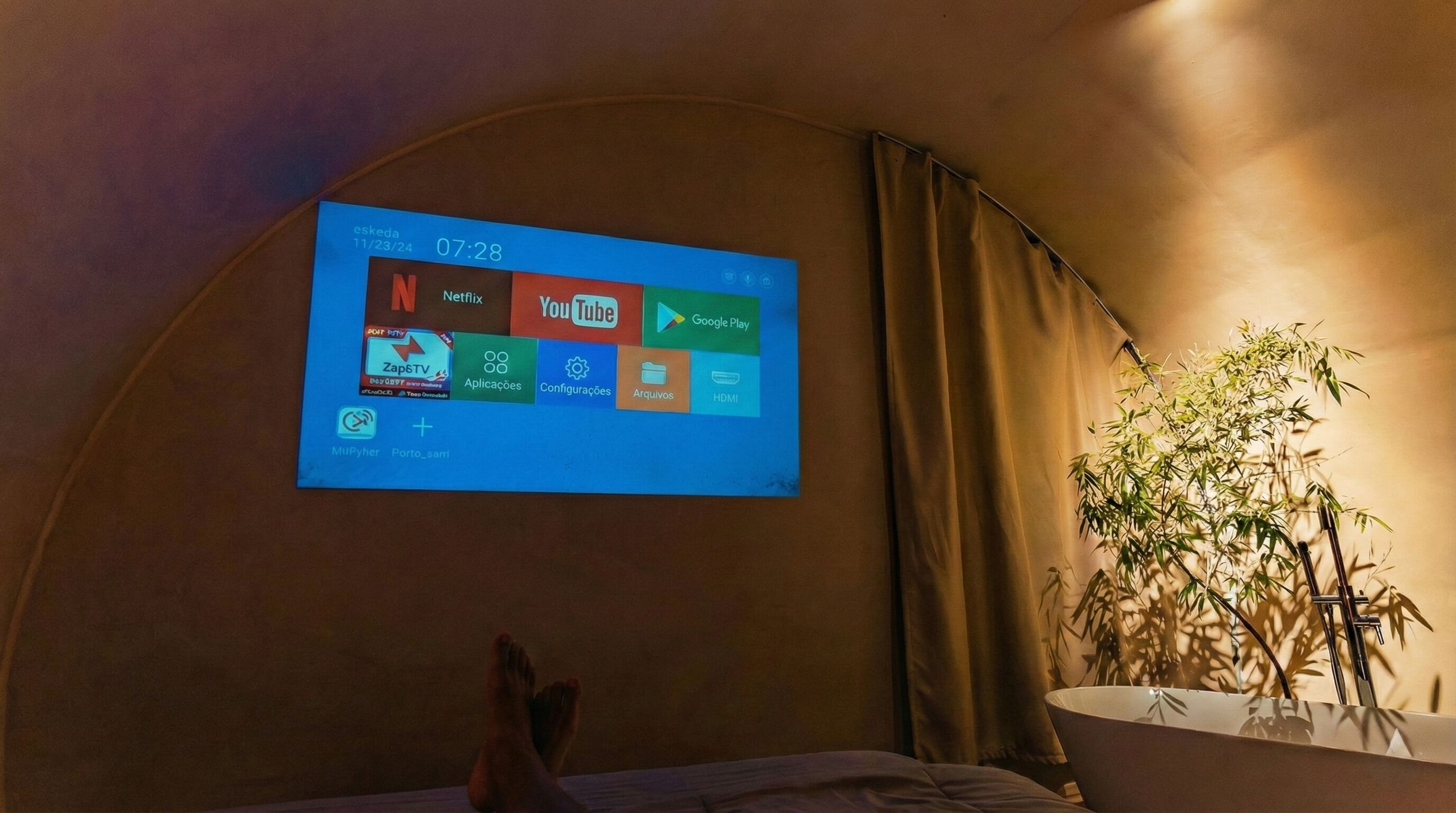 A bed with a person lying down, a wall-mounted screen displaying app icons, and a bamboo plant in the corner.