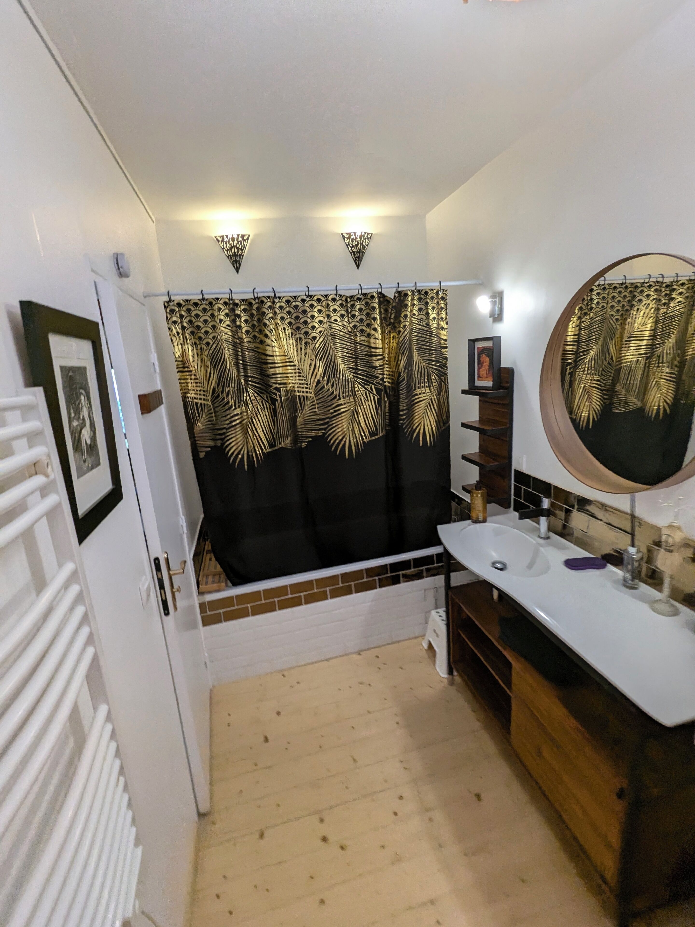 Une salle de bain avec un rideau de douche orné d’un motif de feuilles, un lavabo blanc avec un miroir et un petit tabouret.