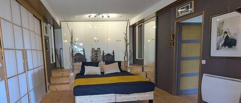 A bedroom with a bed, wooden flooring, a mirror, and a door with a picture frame.