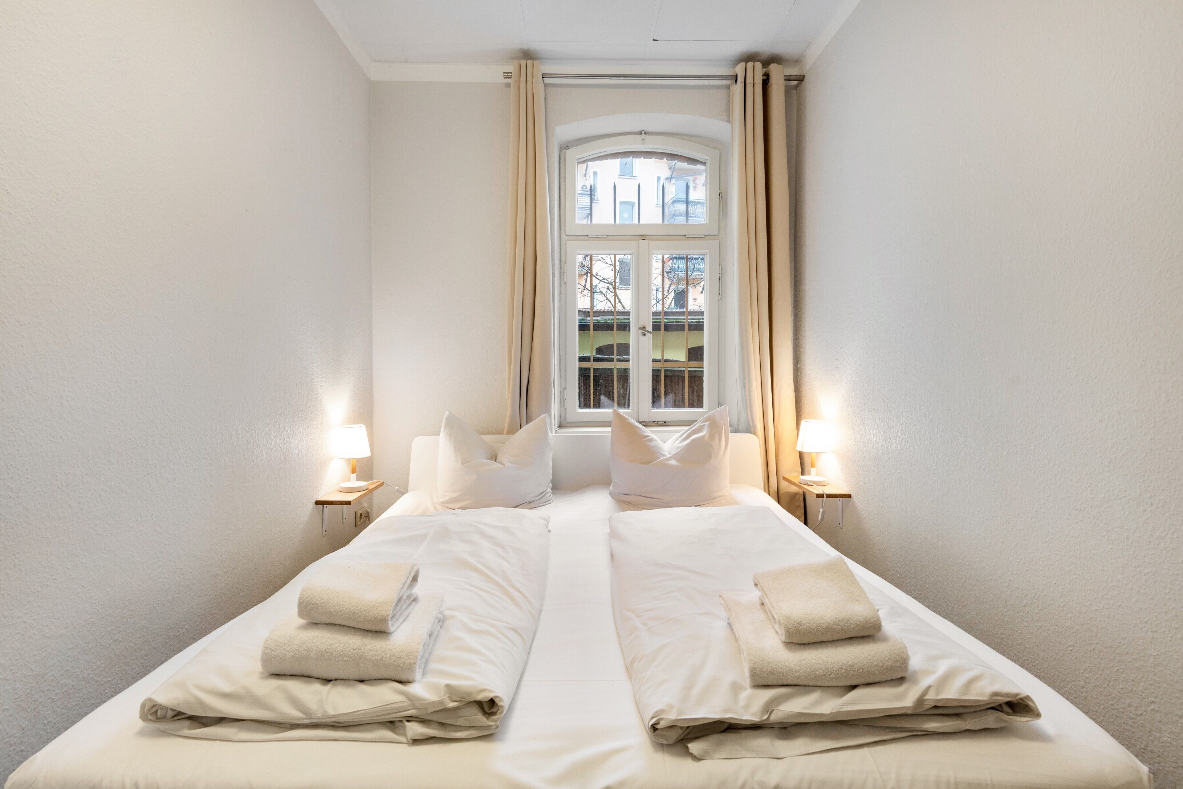 A double bed with white linens and pillows, two bedside lamps, and a window with curtains.