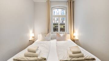 A double bed with white linens and pillows, two bedside lamps, and a window with curtains.