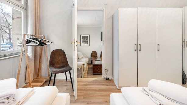 Camera d'albergo con un letto, una sedia, un armadio, uno stendibiancheria e vista attraverso una porta aperta.