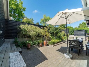 A patio area with a wooden bench, a table with chairs, and a large umbrella. There are potted plants and a wooden fence in the background. - 4 Star Holiday Home in Allinge (Allinge)