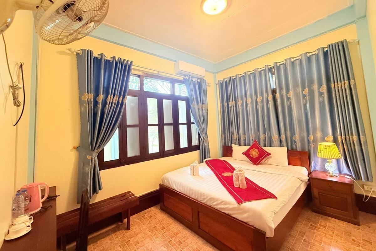 A hotel room with a bed, bedside table, lamp, curtains, and a fan.