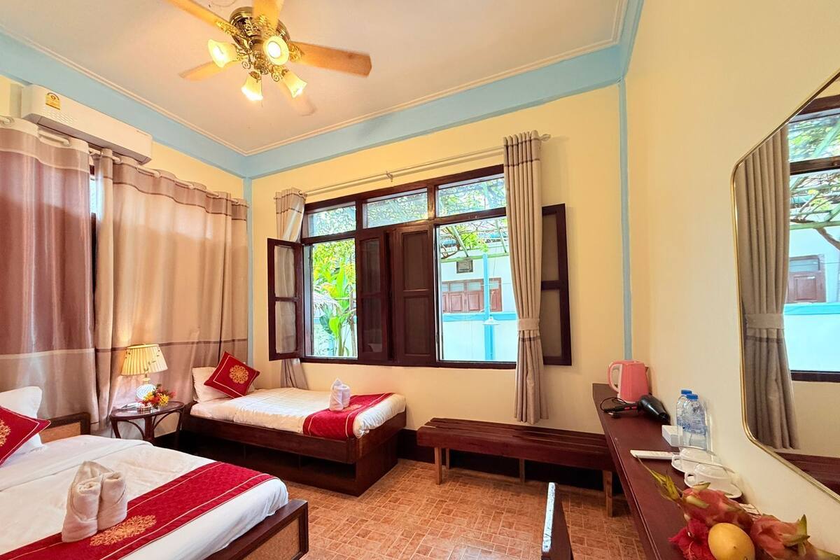 A hotel room with two beds, a desk, a ceiling fan, and large windows with curtains.
