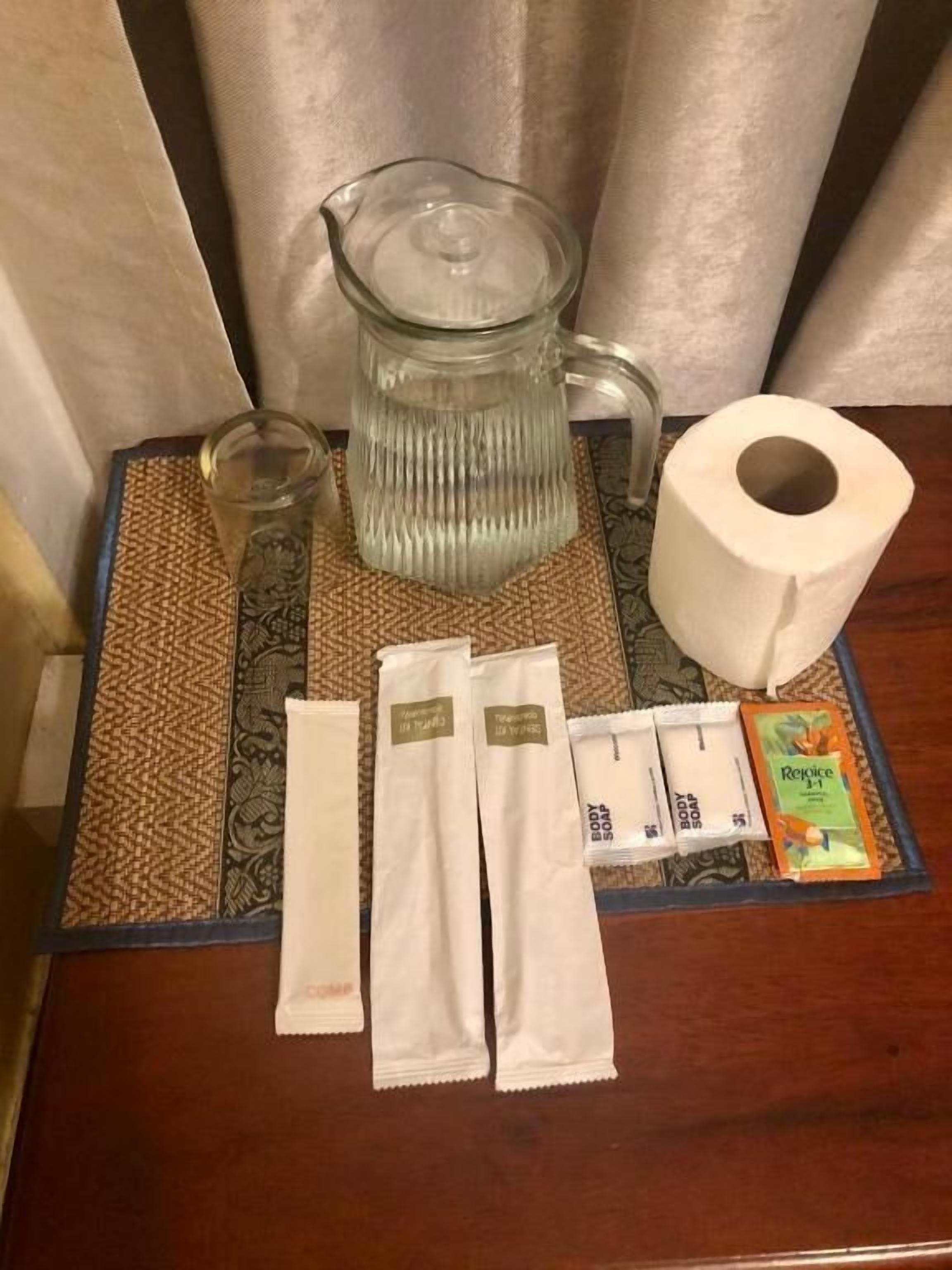 Quarto de hotel com uma mesinha de madeira. Sobre a mesa, há um jarro de vidro, um copo, um rolo de papel higiénico, três saquetas de sabonete líquido e uma saqueta de loção.