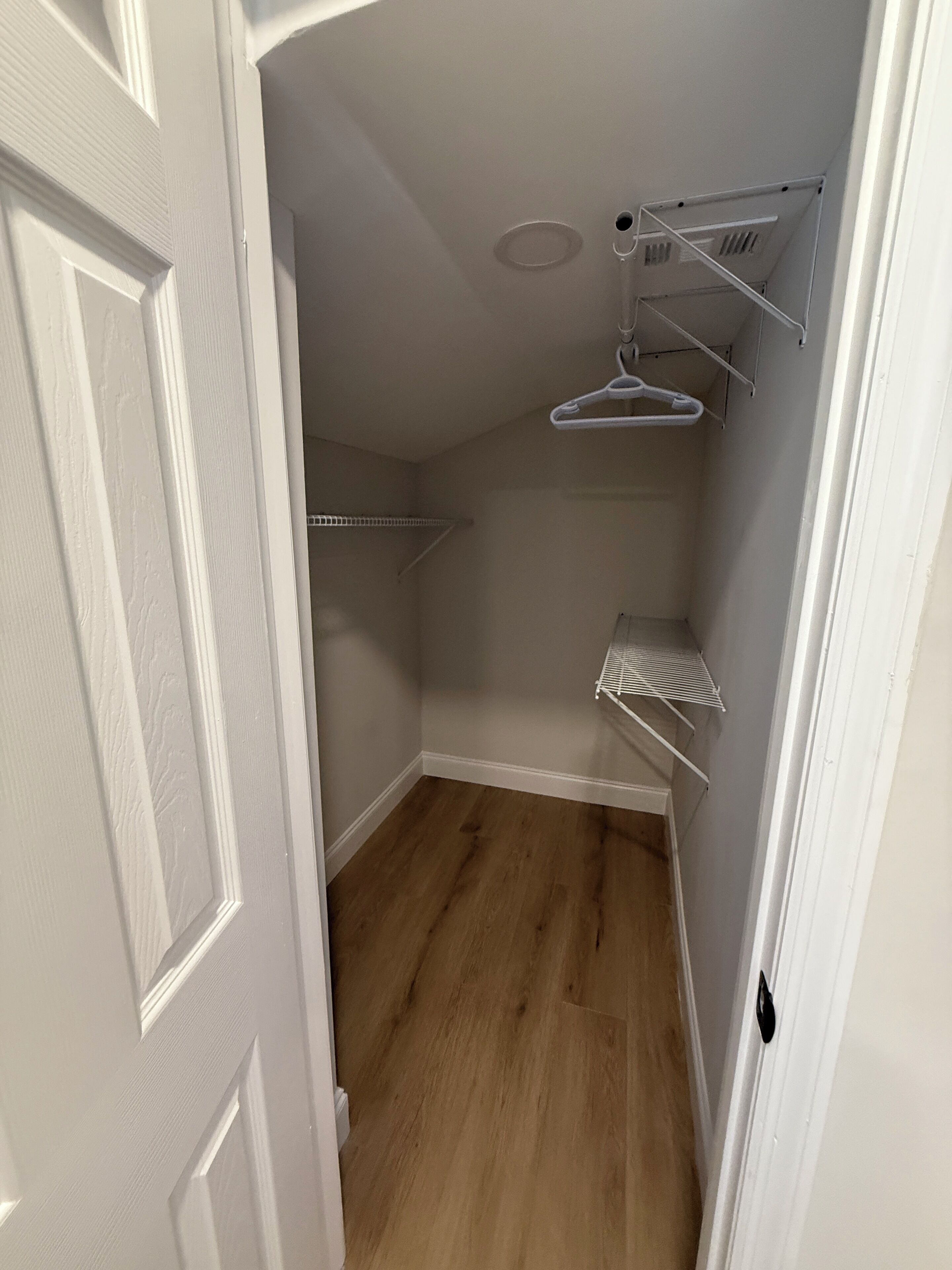 Une petite pièce étroite avec un plancher en bois, des murs blancs et une porte blanche. On y trouve une étagère en métal avec un cintre et un porte-serviettes blanc.