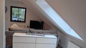 A bedroom with a sloped ceiling, a large window, a bed, a dresser, and a television.
