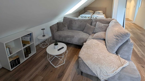 A modern loft bedroom with a sloped ceiling, a large gray sectional sofa, a small white round table, and a built-in bookshelf.