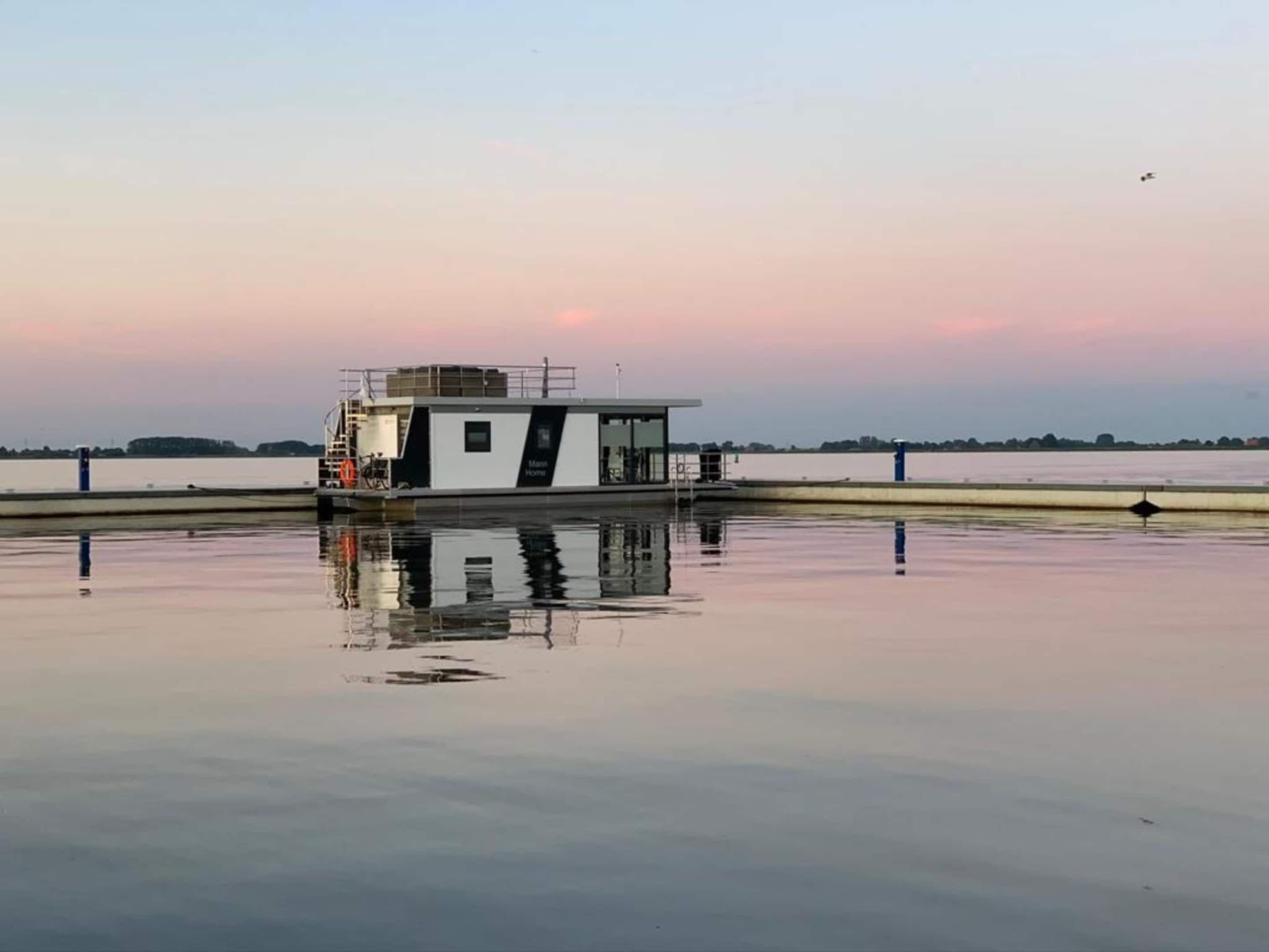 A stationary houseboat with a covered deck and a small cabin, situated on calm water with a clear sky in the background.