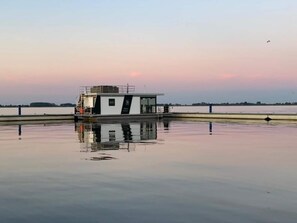 Ein feststehendes Hausboot mit überdachter Terrasse und kleinem Wohnteil, gelegen auf ruhigem Wasser vor einem wolkenlosen Himmel.