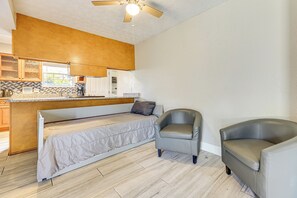 Een compacte kamer met een bed, twee stoelen, een kitchenette en een plafondventilator.