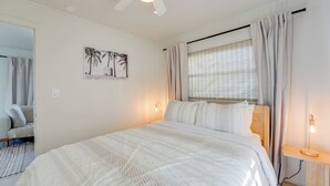 A neatly arranged bedroom with a large bed, a ceiling fan, a window with curtains, a bedside table with a lamp, and a wall-mounted picture. - Beachfront Bliss + Bbqs! Indian Rocks Beach Escape (Indian Rocks Beach)
