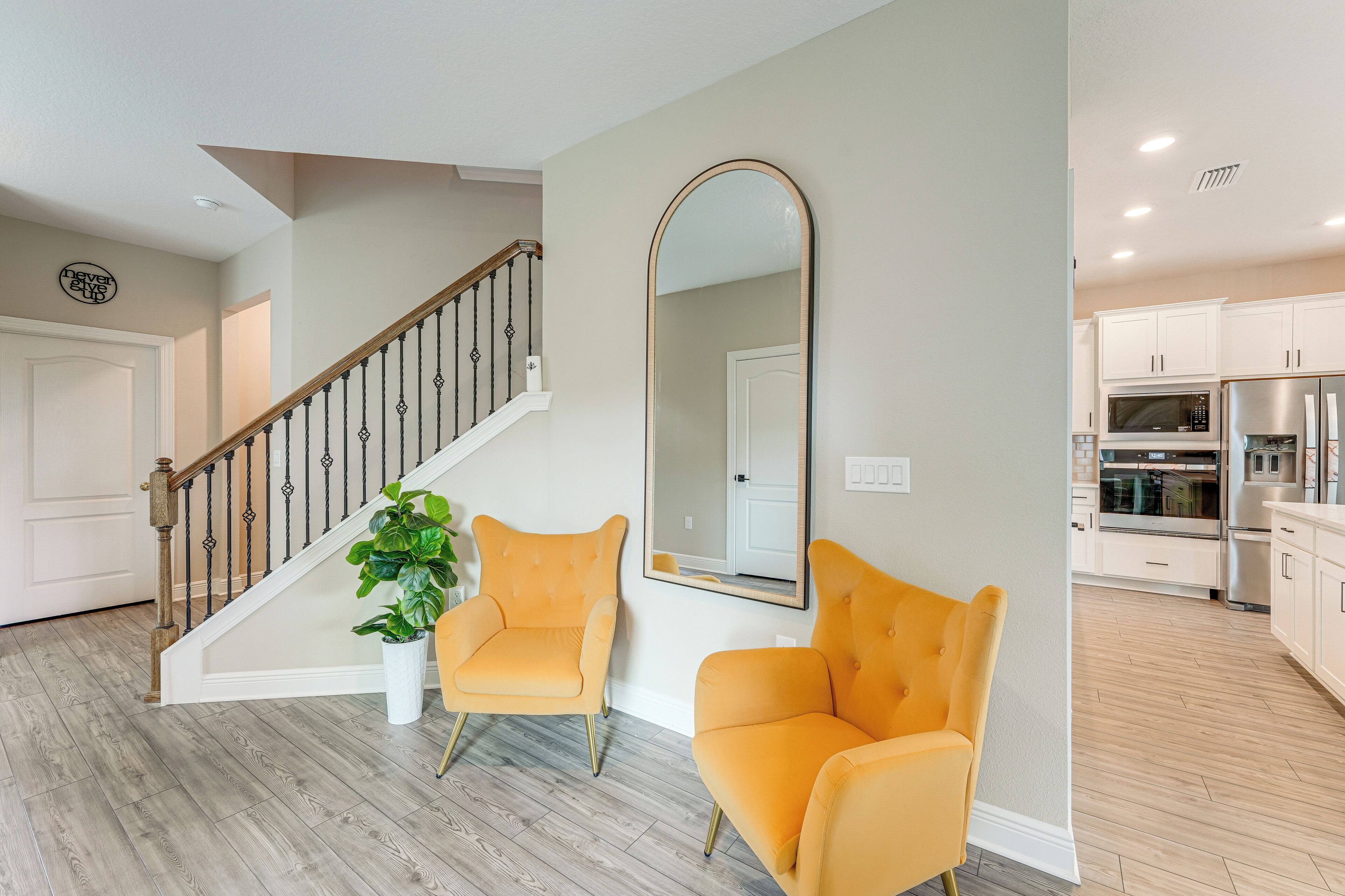 Un salon moderne avec un escalier, deux fauteuils jaunes, un grand miroir et une cuisine en arrière-plan.