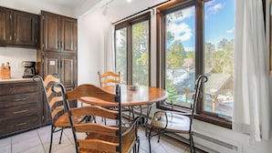 A kitchen with wooden cabinets, a dining table with chairs, and large windows with a view of trees and buildings. - Inner Circle Retreat — Walk to Main Street, Pool and the Slopes (Breckenridge)