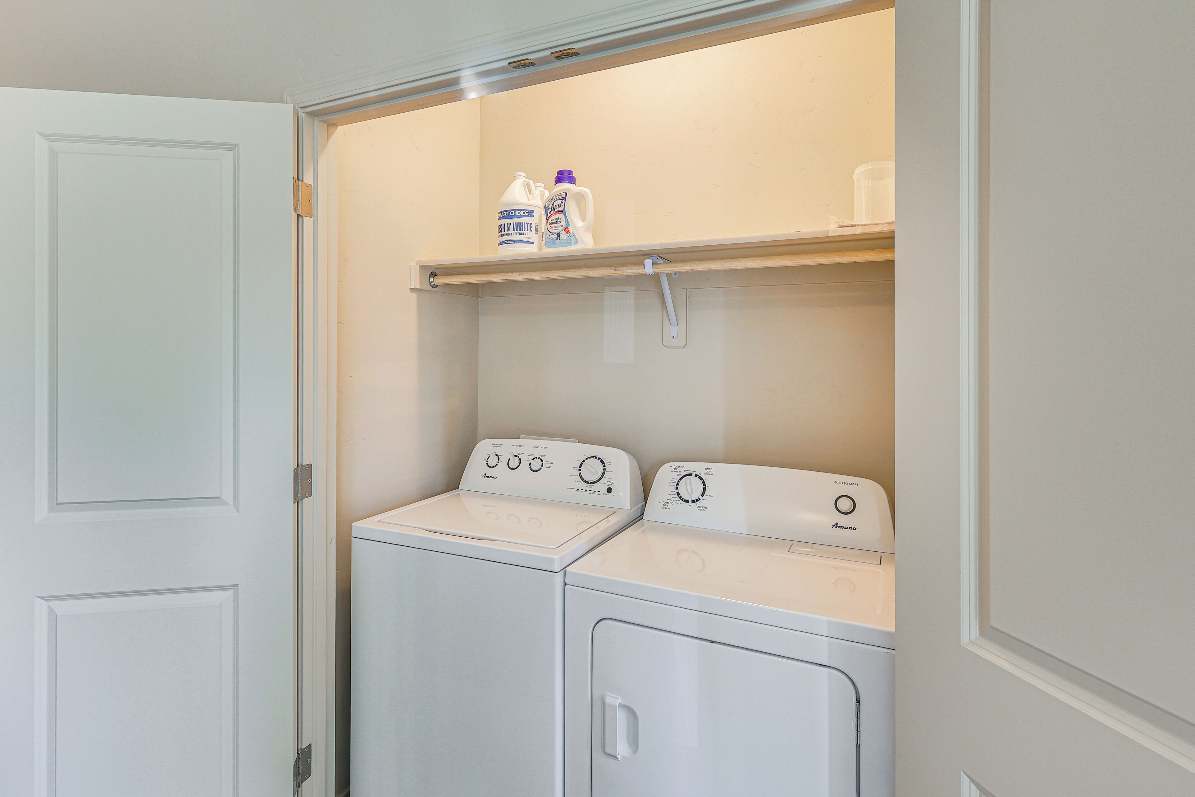 A laundry room with two white front-loading washing machines and a shelf above containing cleaning products.