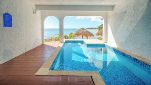 A swimming pool with a view of the ocean, a beach, and a thatched umbrella.