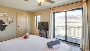 Un dormitorio con cama, televisor, ventilador de techo, puerta y una ventana grande con vistas a las montañas.