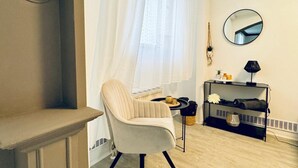 A room with a chair, a small table with a lamp, a shelf with items, and a window with curtains.