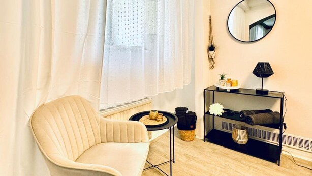 A room with a chair, a small table, a mirror, a shelf with items, and a window with curtains.