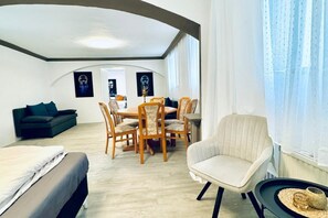A modern room with a bed, a sofa, a dining table with chairs, and a window with sheer curtains.