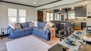 A modern living room with a sectional sofa, a dining table set for six, and a kitchen area with dark cabinets and stainless steel appliances.