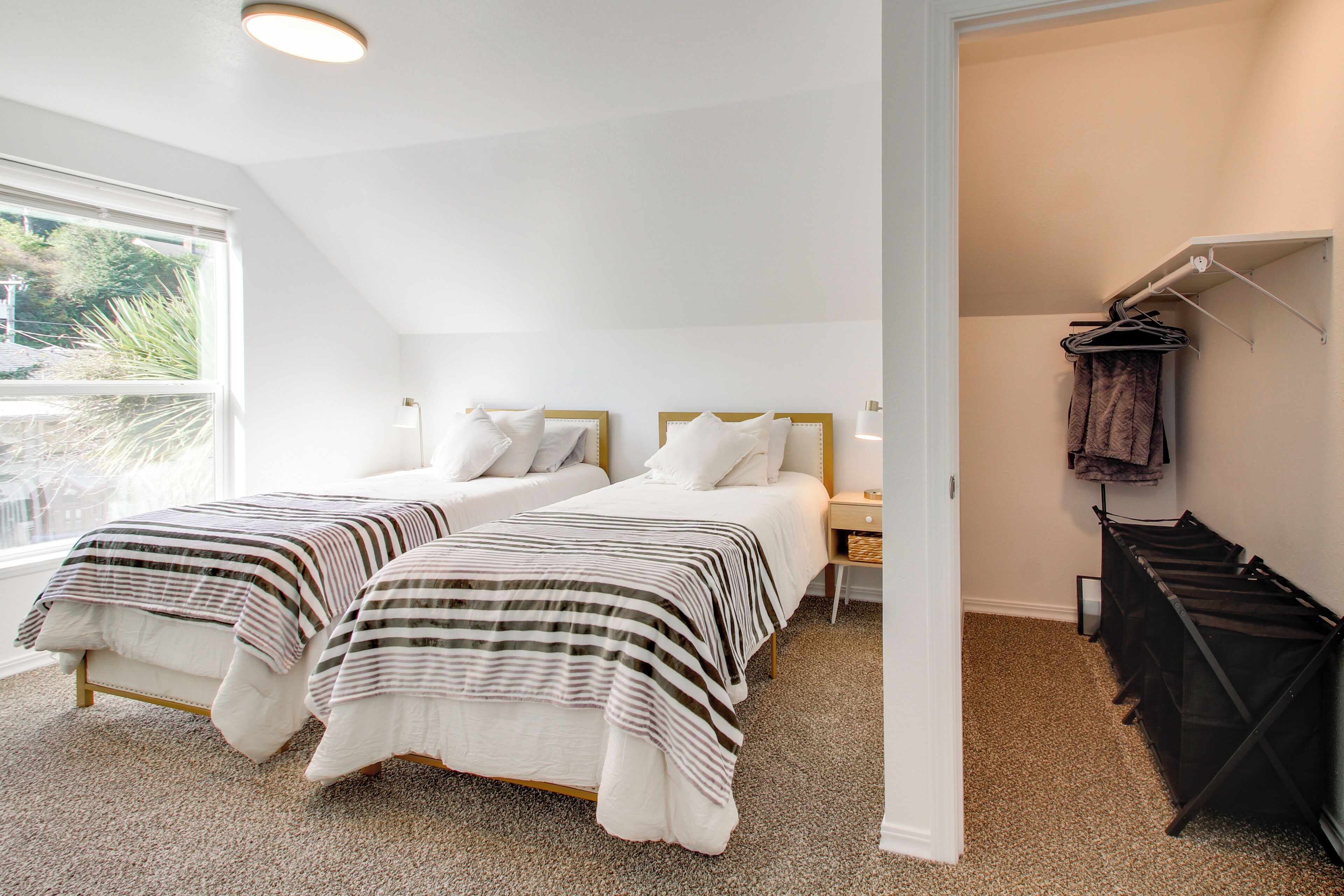 A bedroom with two beds, a window, a closet with hanging clothes, and a carpeted floor.