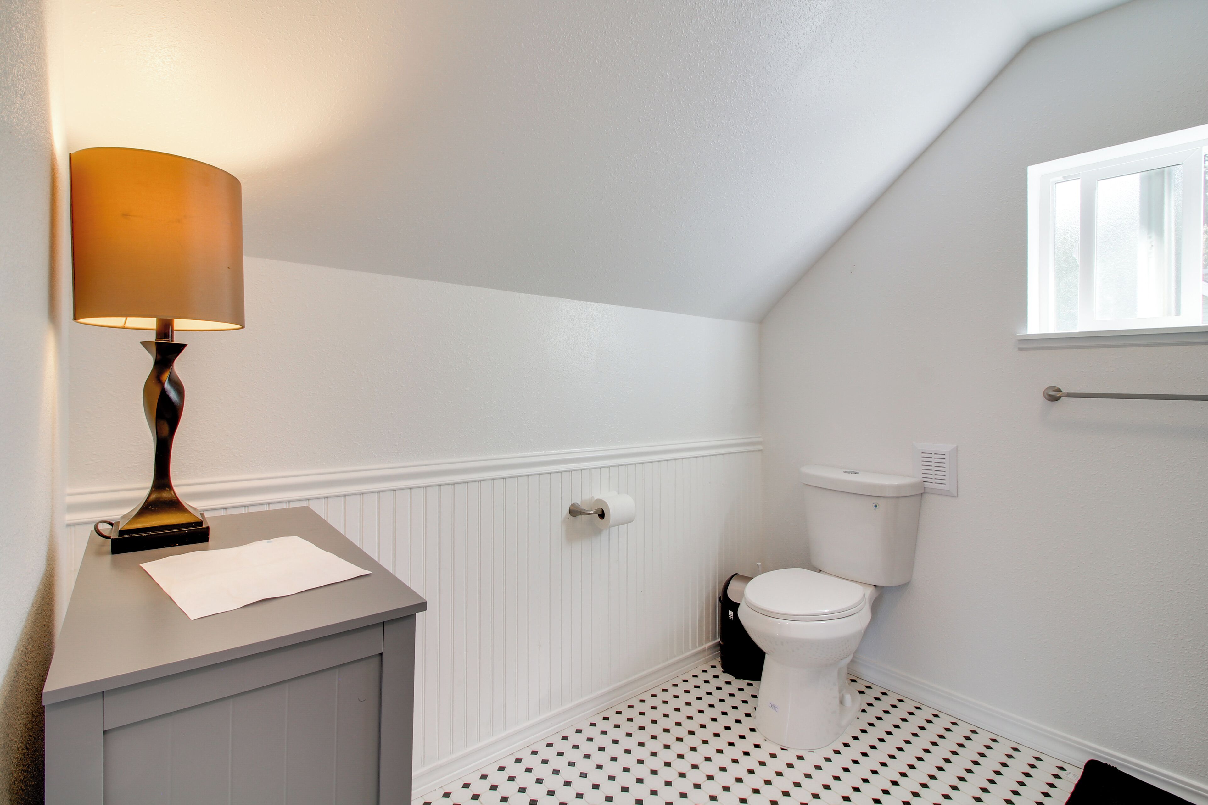 Une salle de bain avec des toilettes blanches, une lampe fixée au mur et une fenêtre.