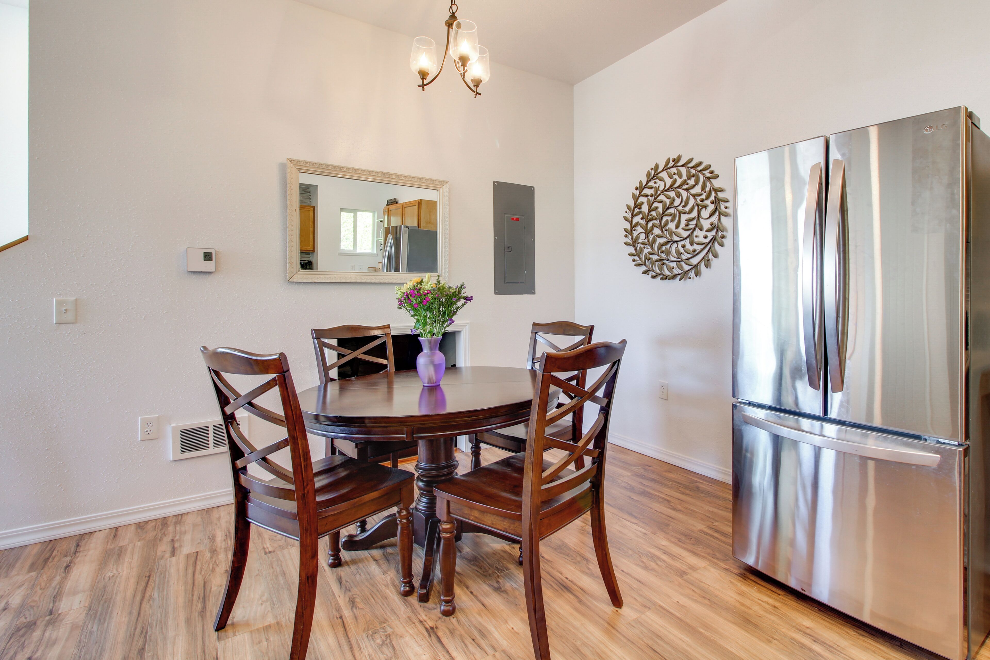 A dining area with a round table, four chairs, a vase with flowers, a chandelier, a refrigerator, and a wall-mounted mirror.