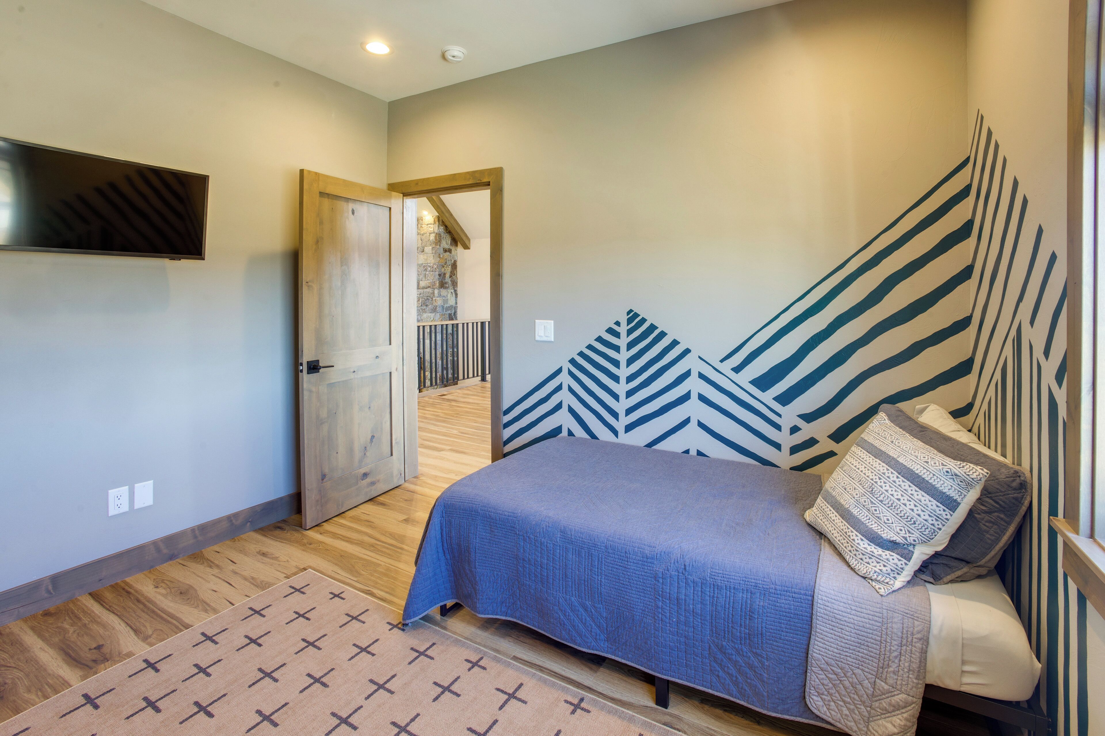 A bedroom with a bed, a TV mounted on the wall, a patterned rug, and a door leading to another room.