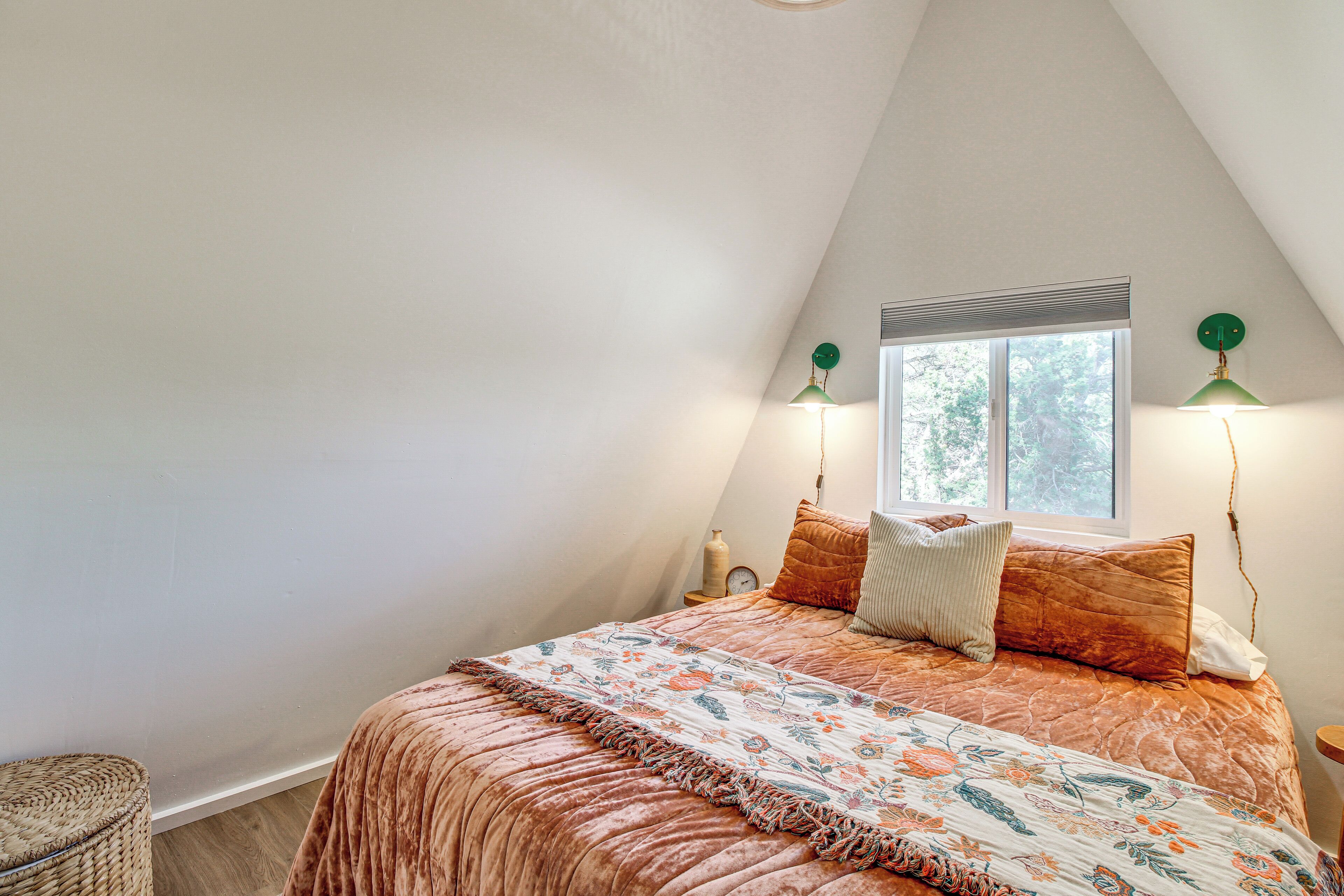 Un dormitorio con techo inclinado, una cama grande con colcha estampada, dos lámparas de noche, una ventana con persianas y una mesita de noche con un reloj.