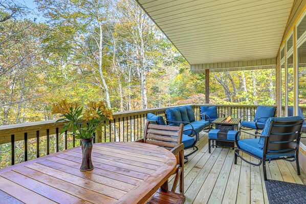 A wooden deck with a table and chairs, a fire pit, and a view of autumn trees.