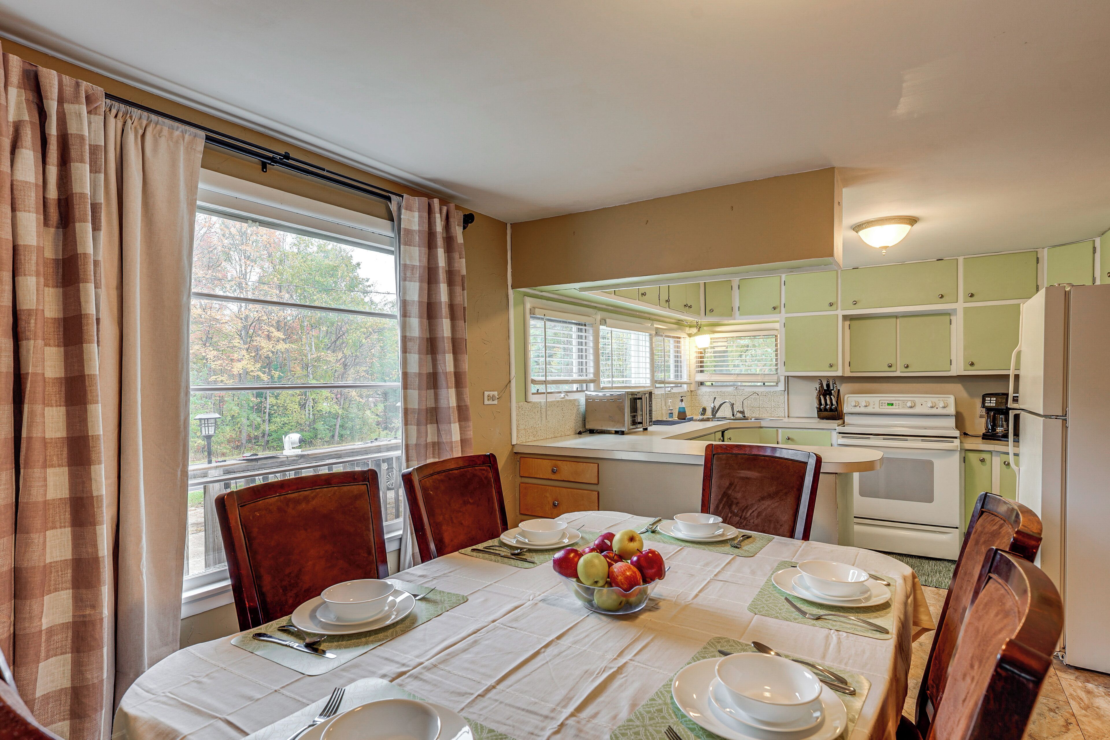 A dining area with a table set for four, a bowl of fruit, and a view of the kitchen in the background.