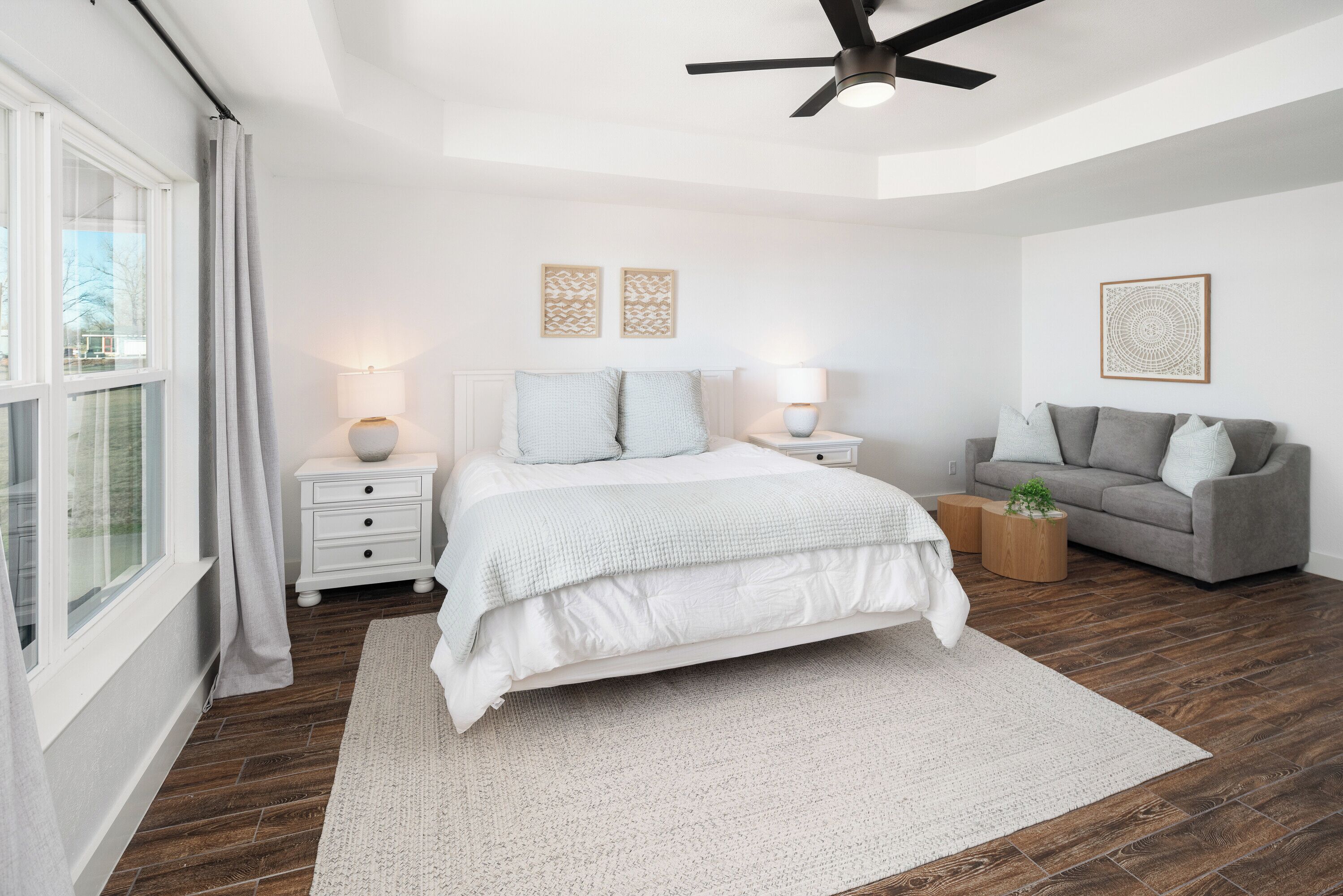 A bedroom with a bed, bedside tables, a grey sofa, a ceiling fan, and a window with curtains.