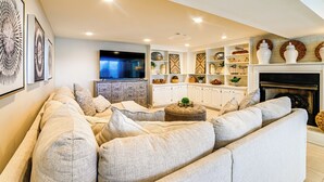 A modern living room with a large sectional sofa, a flat-screen TV, a fireplace, and a kitchen area with white cabinetry and decorative items. - Spacious Kentucky Lake Escape w/ Dock & Views (Benton)