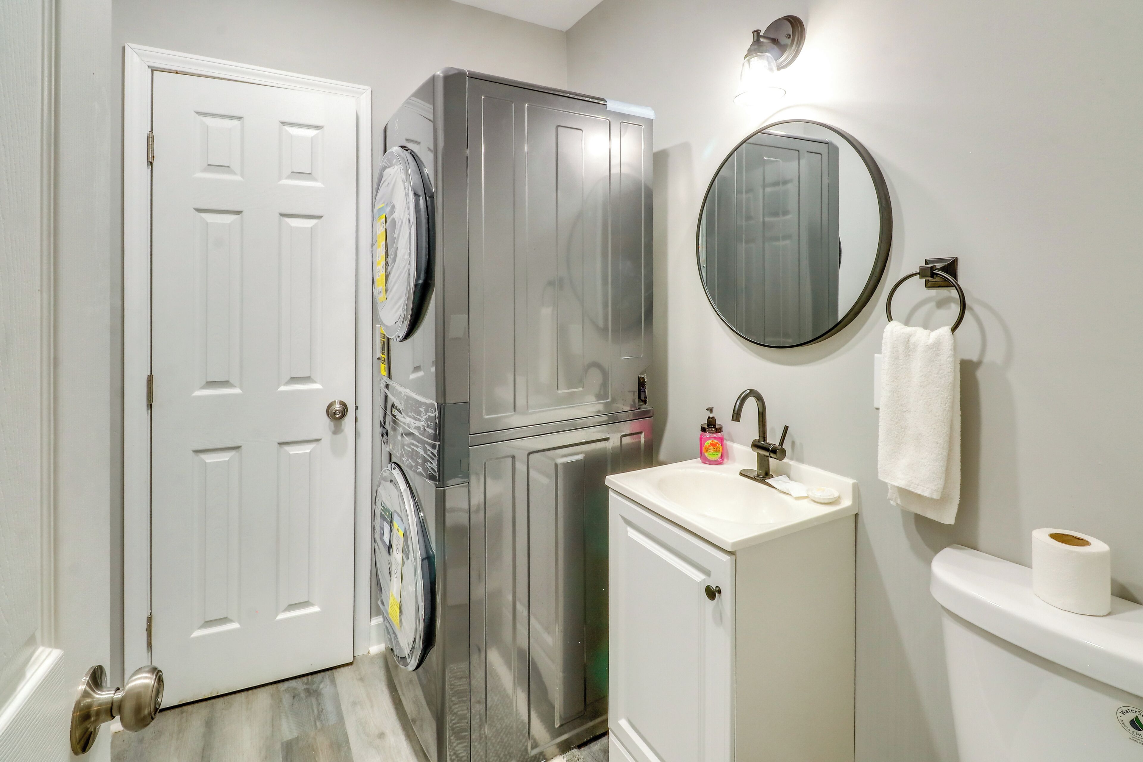 Un baño con una puerta blanca, una lavadora de acero inoxidable, un lavamanos con espejo y un inodoro.